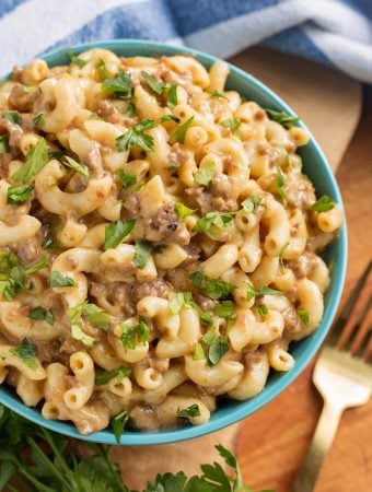 Cheeseburger Macaroni in a blue bowl next to a gold fork all on a wooden board