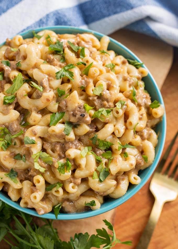 Cheeseburger Macaroni in a blue bowl next to a gold fork all on a wooden board