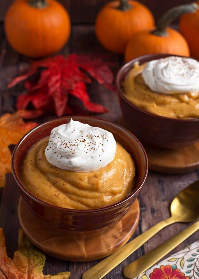 Quick Pumpkin Pie Pudding topped with a dollop of whipped cream on wooden coasters in front of red and orange leaves and pumpkins