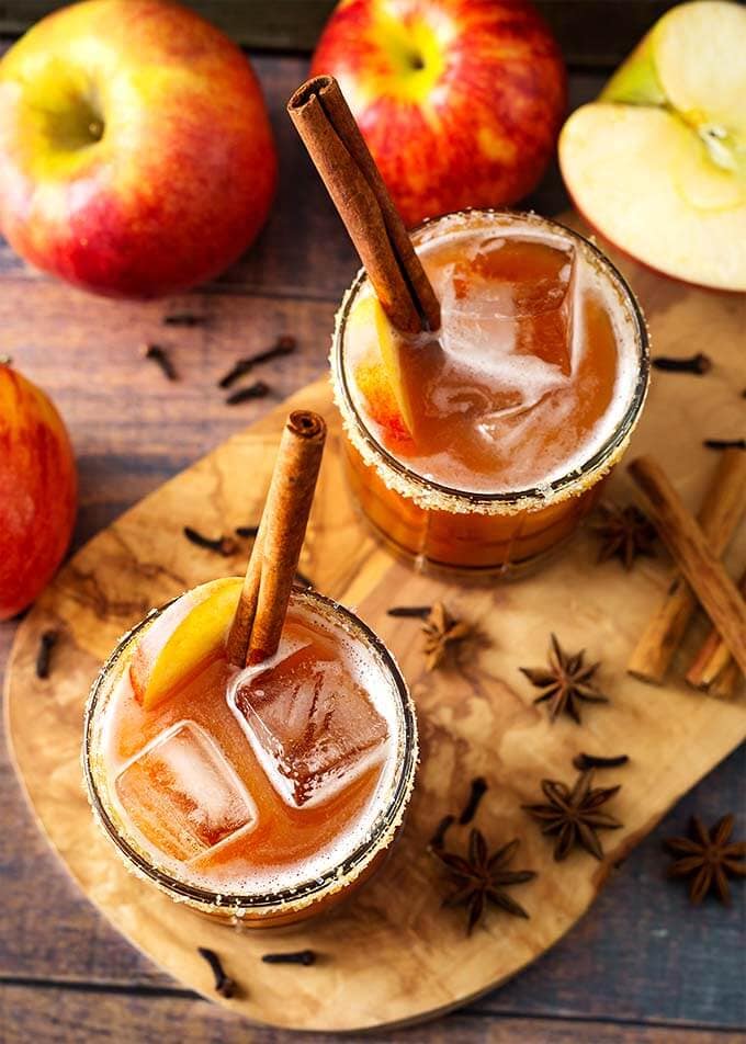 Top view of two Apple Butter Old Fashioned Cocktails with ice cubes, cinnamon sticks, and apple slices all on a wooden board