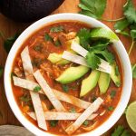 Chicken Tortilla Soup in a white bowl topped with tortilla strips, sliced avocados, and cilantro