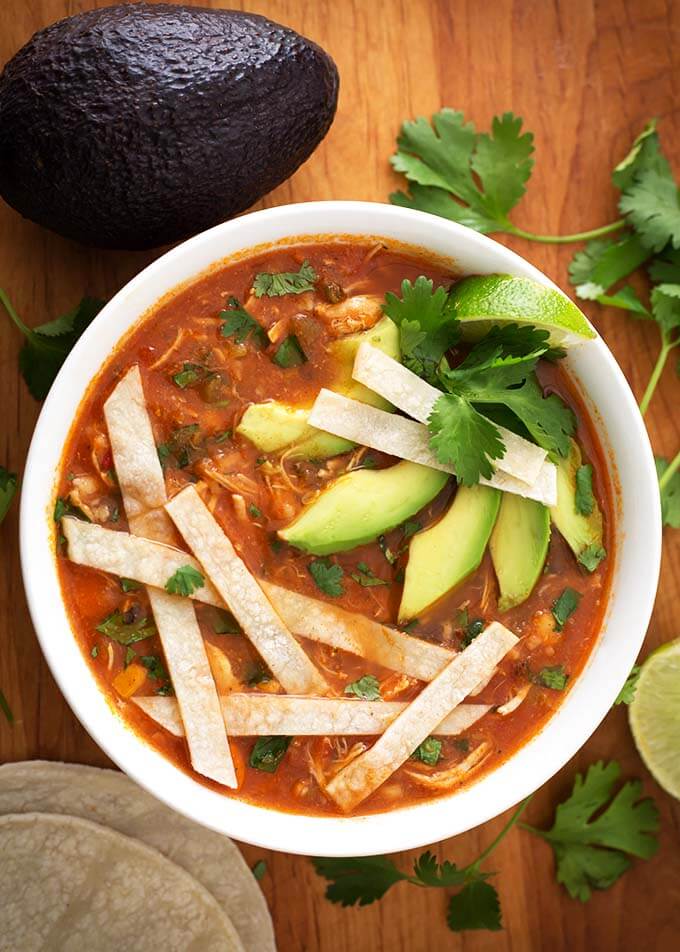 Chicken Tortilla Soup in a white bowl topped with sliced tortilla strips, cilantro, and sliced avocado all on a wooden board