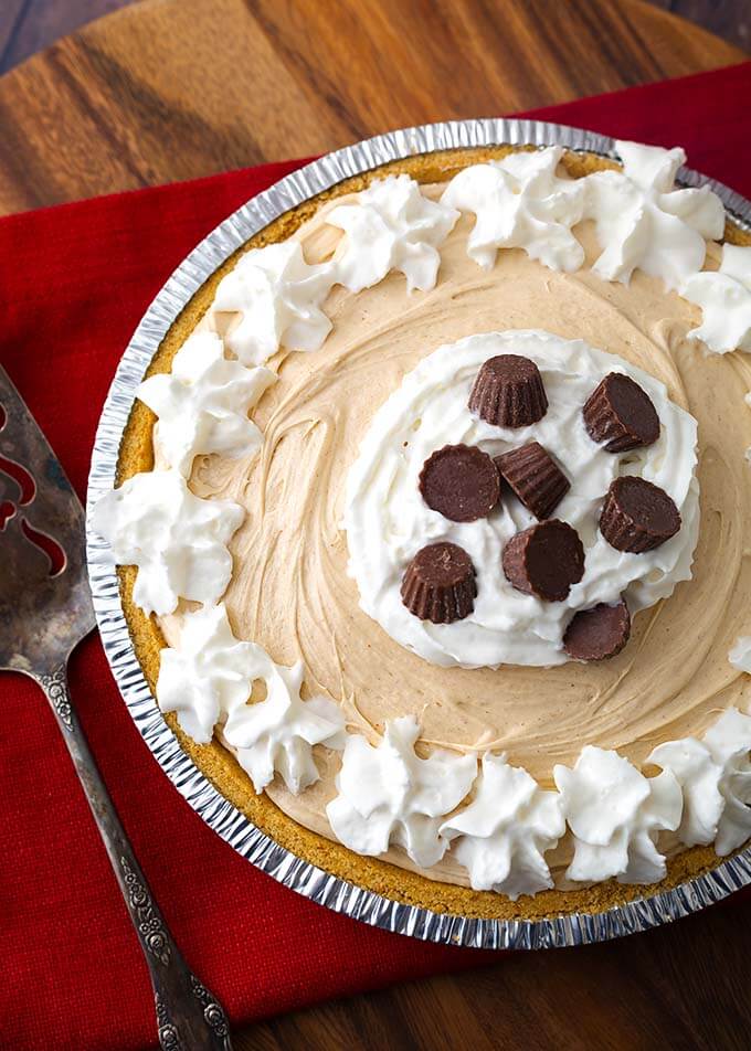 No Bake Peanut Butter Pie topped with whipped cream and chocolate candies in a disposable pie tin next to a silver serving spoon all on a wooden board