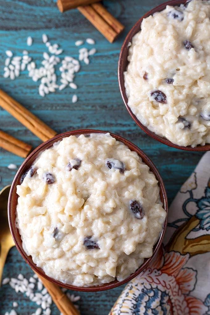 Two brown bowls of Rice Pudding next to cinnamon sticks on a turquoise background