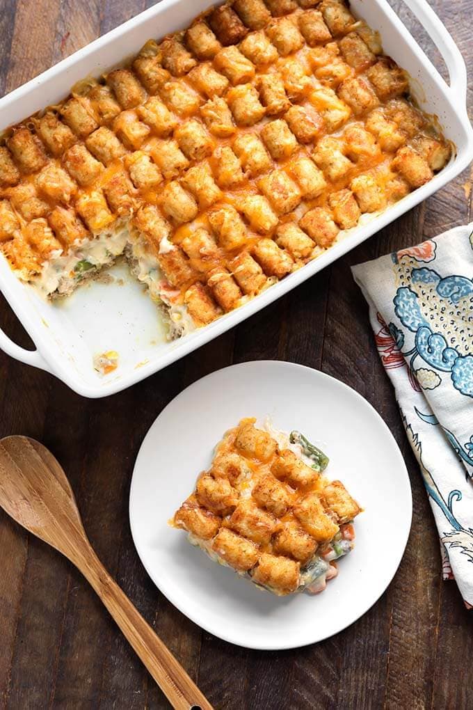 Slice of Hotdish Tater Tot Casserole on a white plate in front of remaining casserole in a baking dish