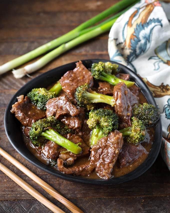 Beef and Broccoli in a black bowl next to two wooden chosticks and a black and white patterned napkin