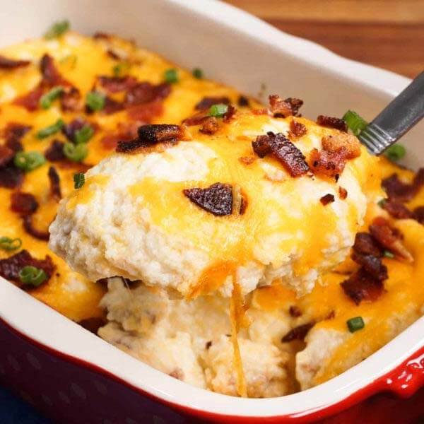 baked cauliflower casserolein a white and red baking dish with serving spoon scooping out a slice