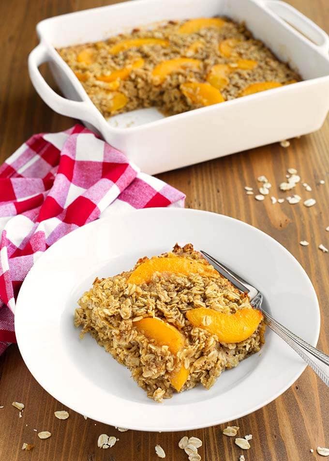 Slice of Easy Peach Baked Oatmeal with a fork on a white plate in front of white baking dish with rest of peach baked oatmeal