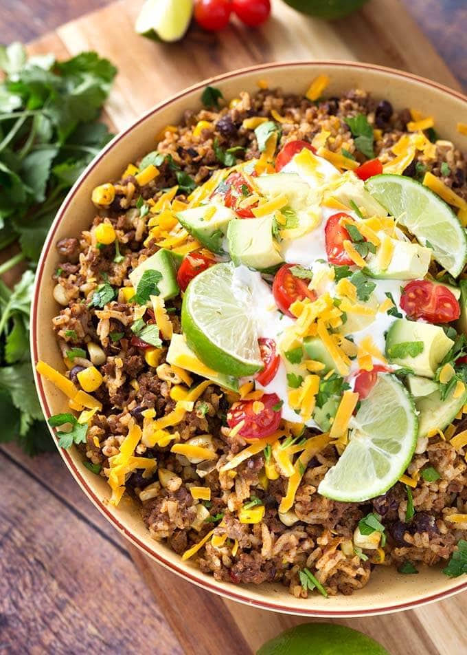 Beef Taco Bowls in a white bowl topped with sliced limes, shredded cheese, diced tomatoes, and sour cream