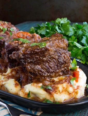 Short Ribs over mashed potatoes on a black plate next to a bunch of fresh parsley