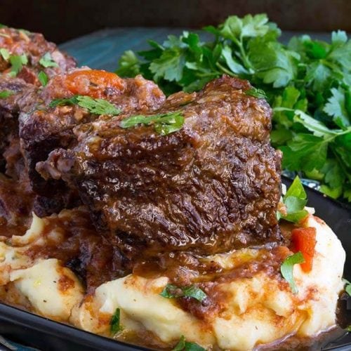 Short Ribs over mashed potatoes on a black plate next to a bunch of fresh parsley