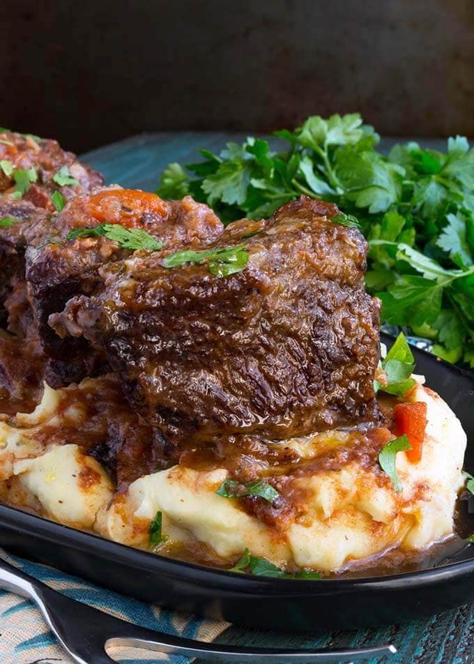 Short Ribs over mashed potatoes on a black plate next to a bunch of fresh parsley