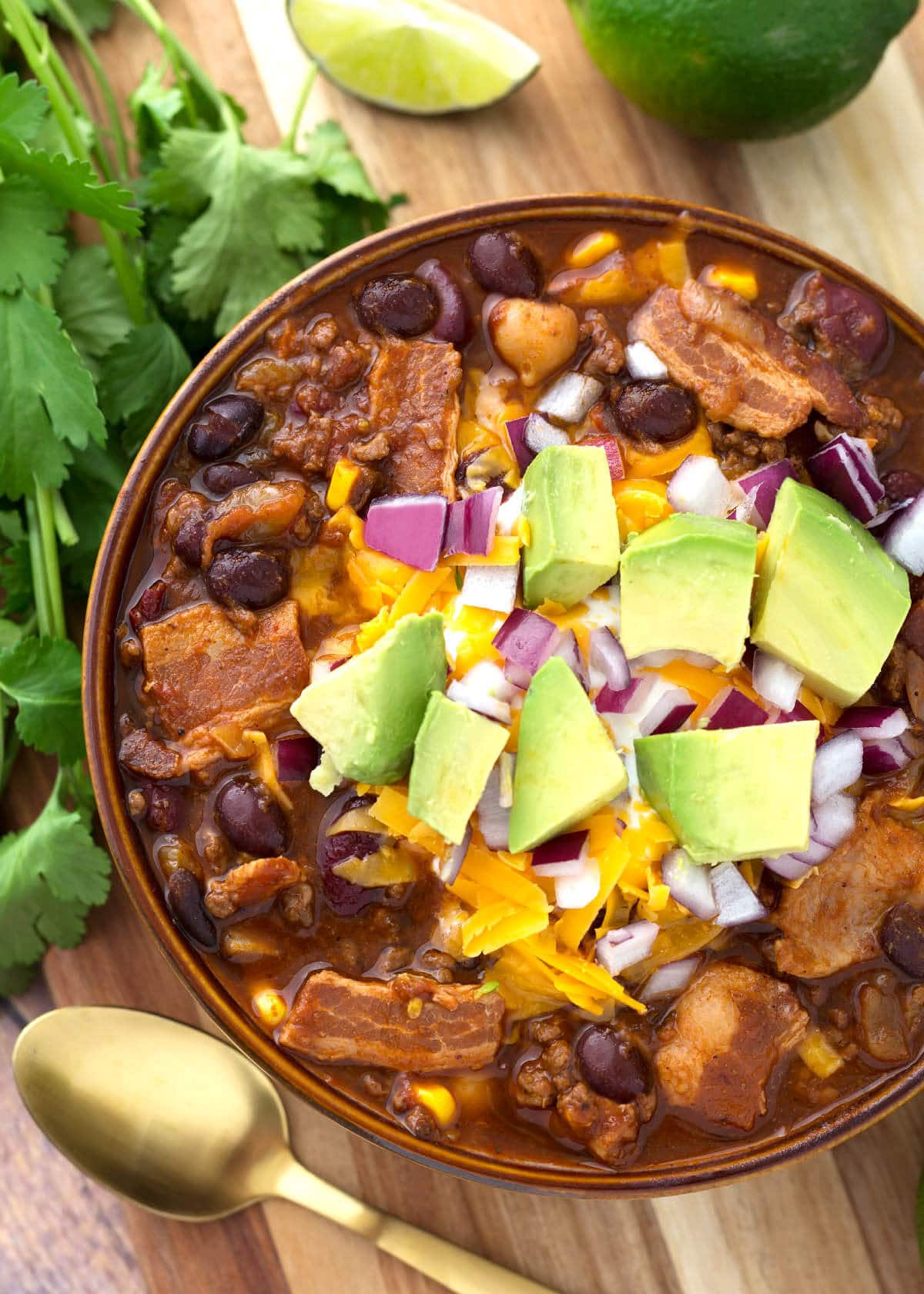 Beefy Bacon Chili in a brown bowl topped with cubed avocado, shredded cheese, and diced red onion