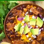 Beefy Bacon Chili in a brown bowl topped with cubed avocado, shredded cheese, and diced red onion