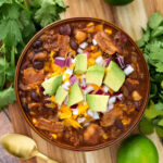 Beefy Bacon Chili in a brown bowl topped with cubed avocado, shredded cheese, and diced red onion