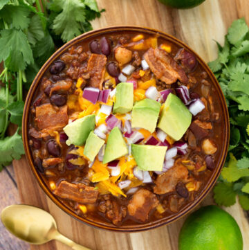 Beefy Bacon Chili in a brown bowl topped with cubed avocado, shredded cheese, and diced red onion