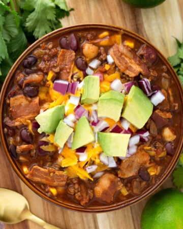 Beefy Bacon Chili in a brown bowl topped with cubed avocado, shredded cheese, and diced red onion