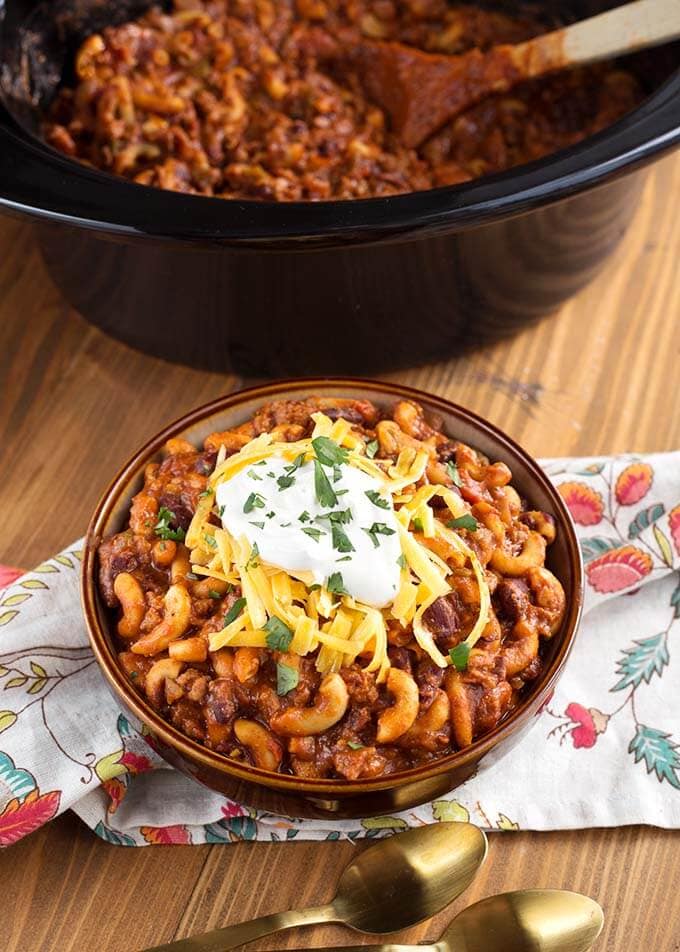 Chili Mac in a brown bowl topped with cheese and sour cream in front of a slow cooker