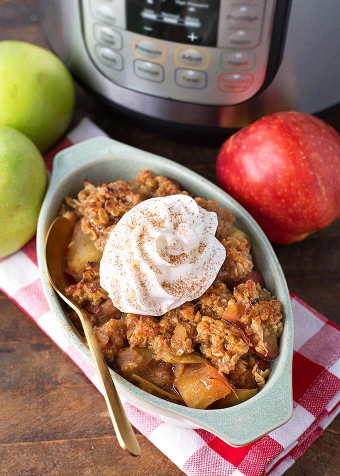 Apple Crisp in oblong gray baking dish topped with whipped cream in front of apples and pressure cooker