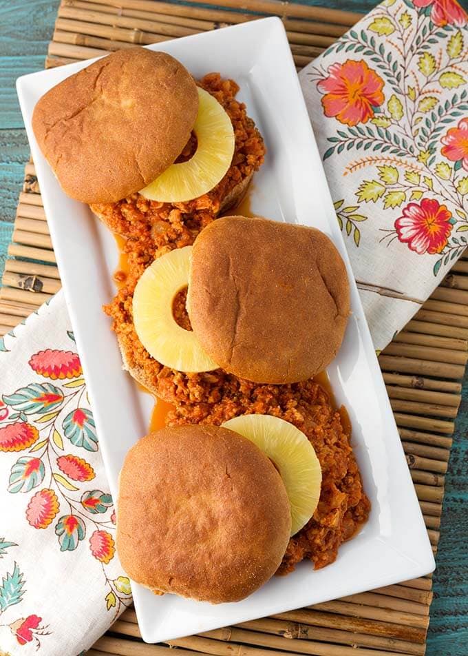 Top view of three Hawaiian BBQ Sloppy Joes  with slices of pineapple on buns on a wite serving platter on top of a floral dish towel