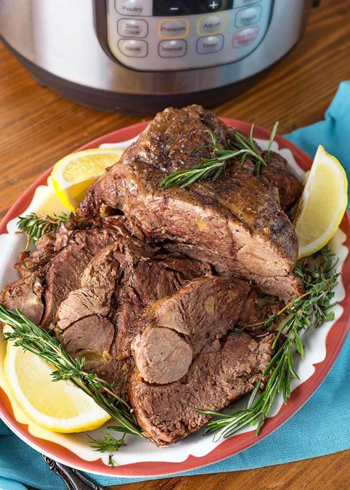 Leg of Lamb garnished with rosemary and lemons on red bordered plate in front of pressure cooker