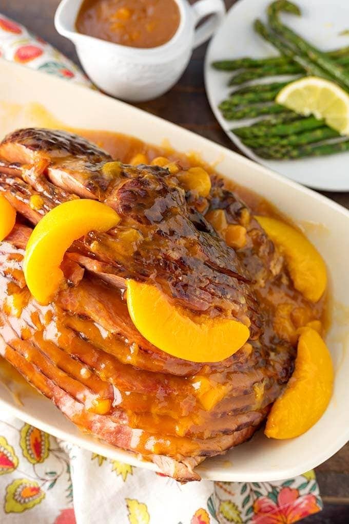 Peach Glazed Ham on white serving platter next to white plate with asparagus and small gravy pitcher