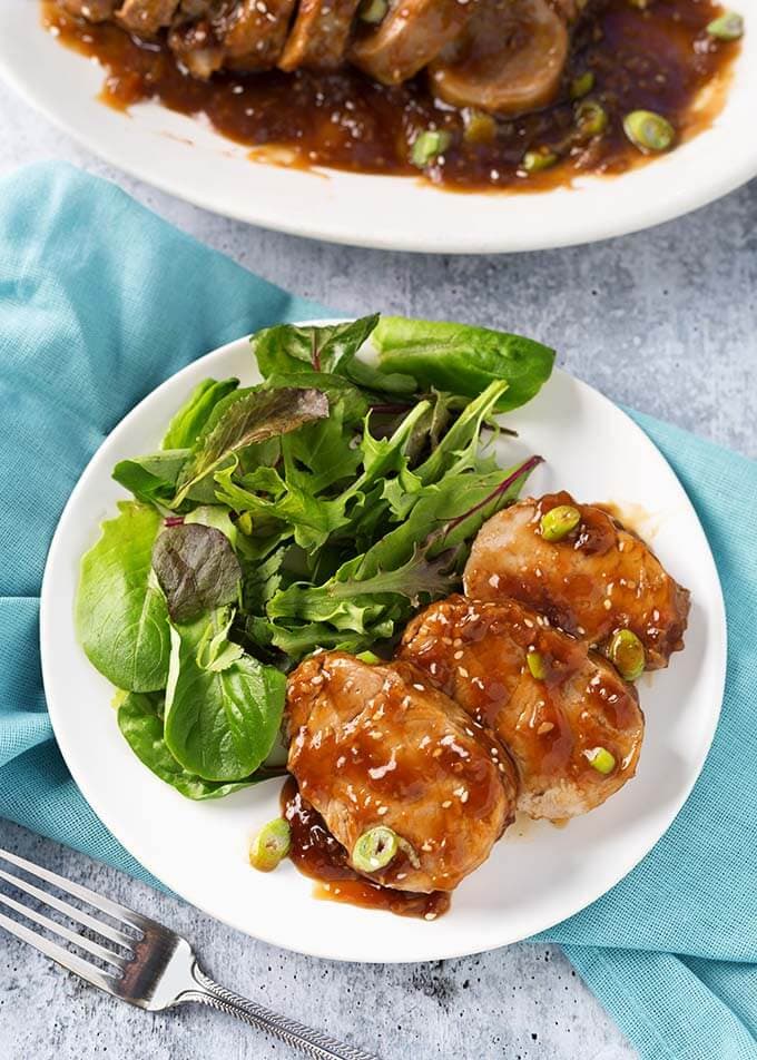 Sliced Teriyaki Pork Tenderloin with green leafy vegetables on a white plate on blue napkin