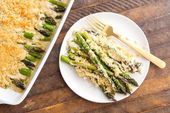 Creamy Baked Asparagus Alfredo on white plate with gold fork next to baking dish of creamy baked asparagus Alfredo