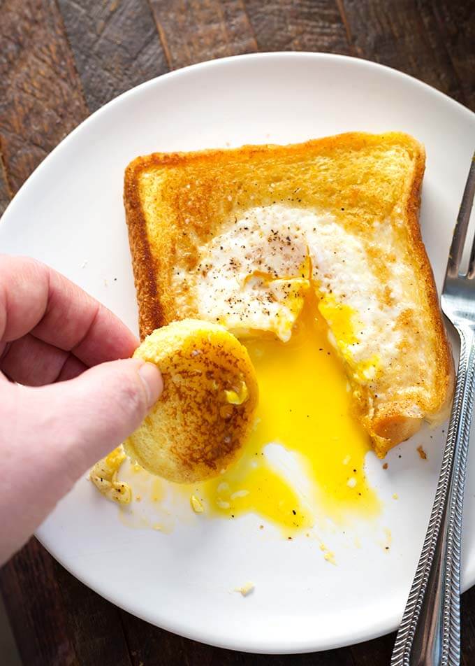 Hand dipping circle toast into Egg in a Hole on a white plate