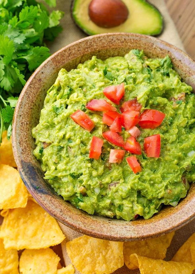 Fresh Homemade Guacamole in brown bowl topped with diced tomatoes next to corn tortilla chips