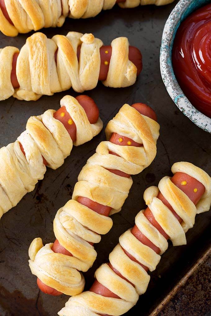 Baked Halloween Hot Dog Mummies on baking sheet with small bowl of ketchup