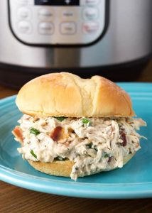 Crack Chicken on hamburger bun on blue plate in front of pressure cooker