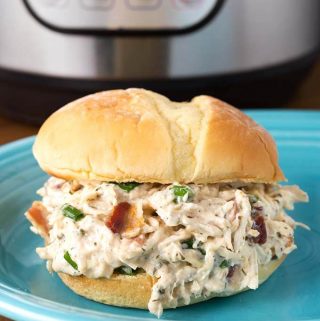 Crack Chicken on hamburger bun on blue plate in front of pressure cooker