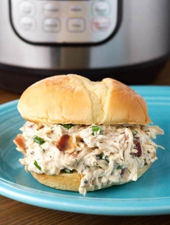 Crack Chicken on hamburger bun on blue plate in front of pressure cooker