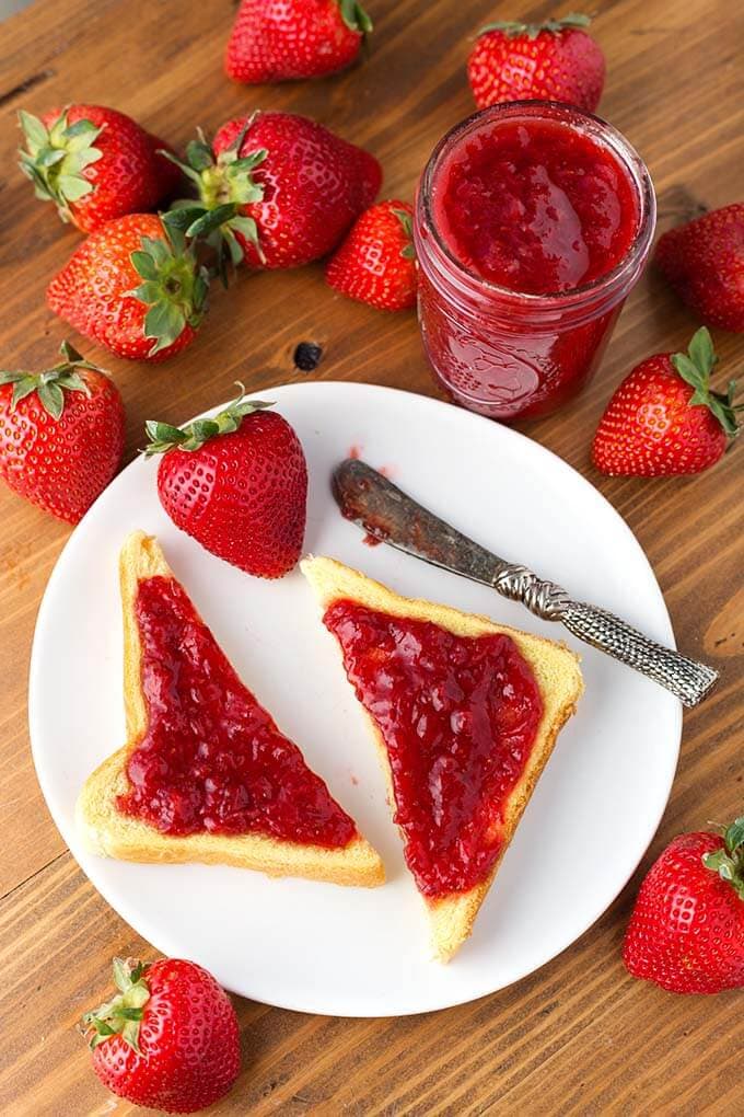 Strawberry Jam spread on cut toast on white plate with strawberries and silver knife in front of jar