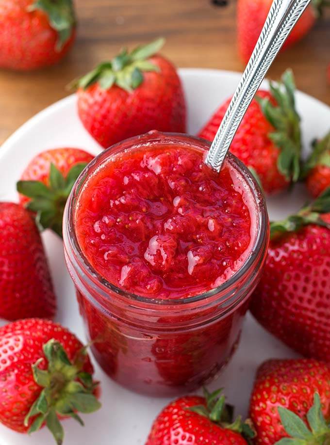 Strawberry Jam in small canning jar with silver spoon on a white plate surrounded by strawberries