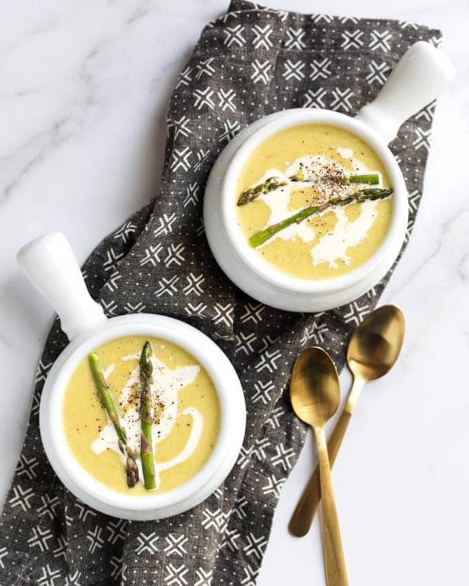 Two white bowls of Asparagus Soup and two golden spoons on a black patterned napkin
