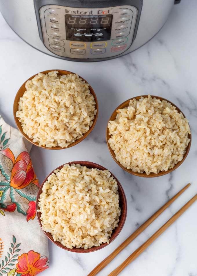 Top view of three bowls of brown rice and chopsticks in front of pressure cooker