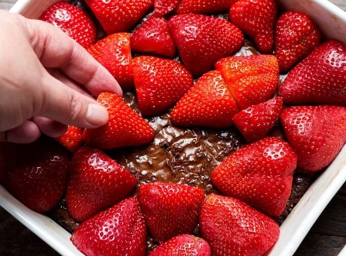 Hand placing strawberries on top of brownie in white baking dish
