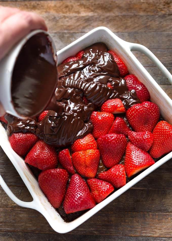 hand pouring chocolate ganache over strawberries and brownie in baking dish