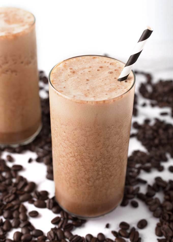 Iced Coffee Protein Shake in glass with brown and white stripped straw surrounded by coffee beans