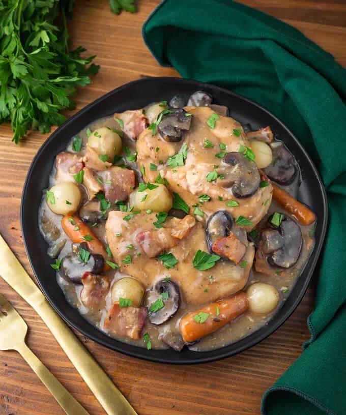 Coq Au Vin in a black bowl on wooden board next to gold fork and knife