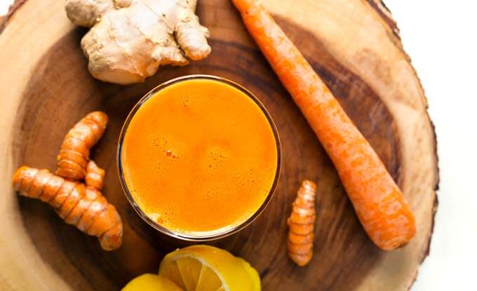 Top view of Fresh Turmeric Tonic surrounded by turmeric root, ginger foot, lemons and a carrot on a wooden board