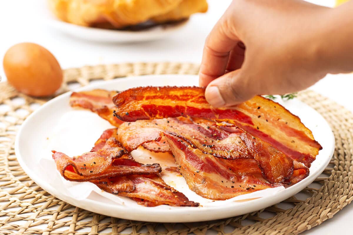 white plate of bacon and hand reaching for a slice