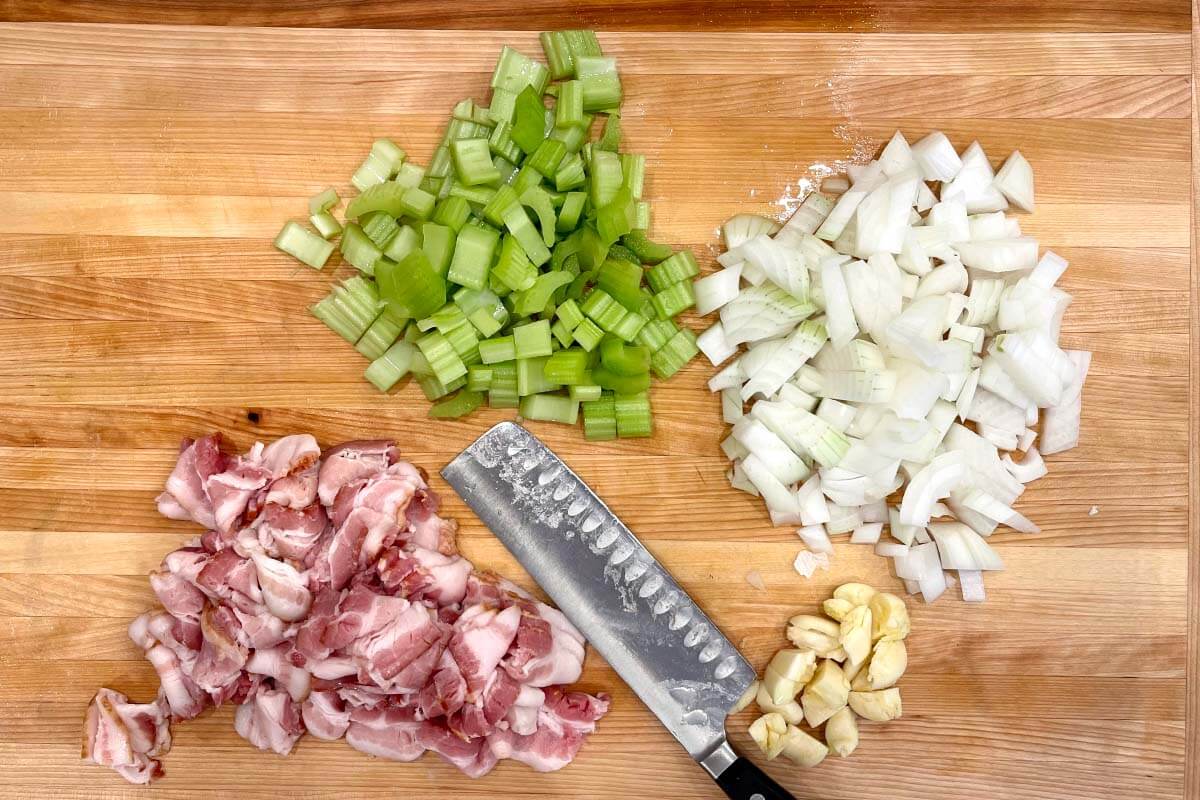bacon, celery, onion, garlic chopped on board with knife.