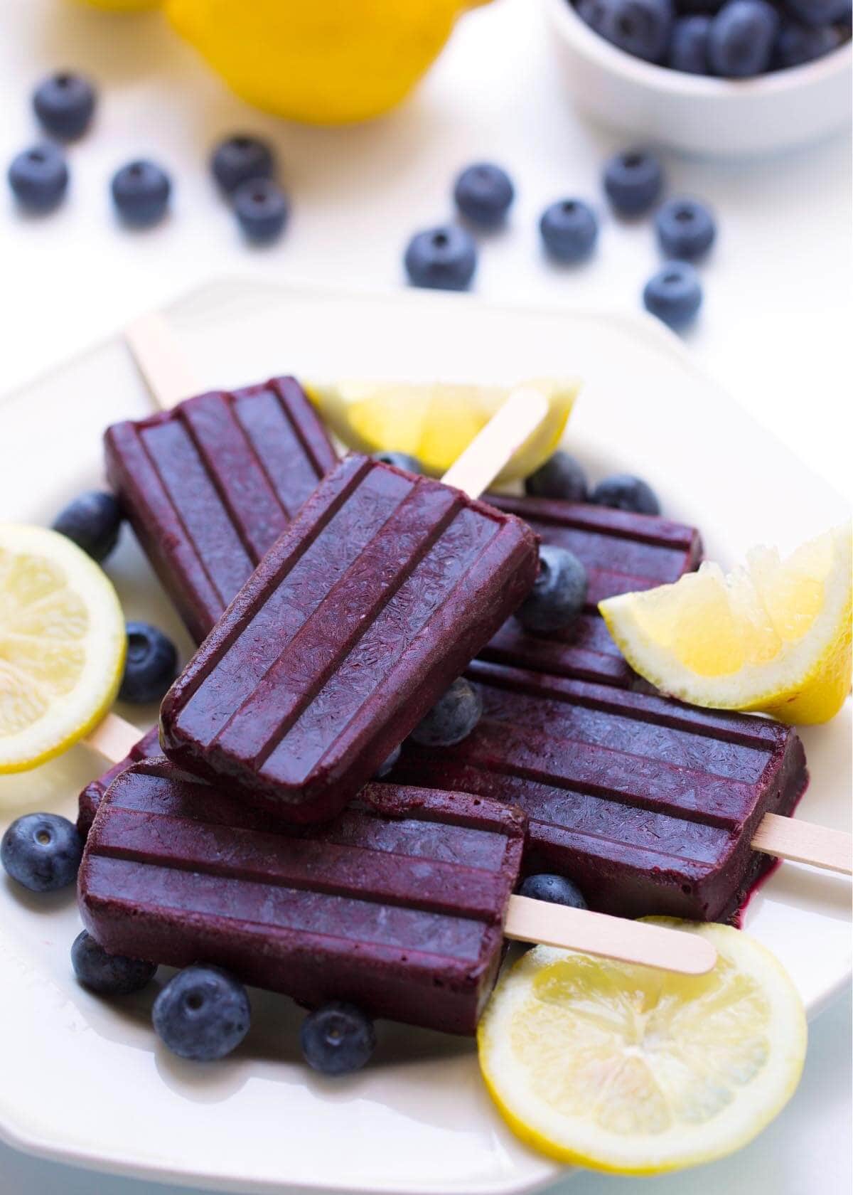 Five Blueberry Lemon Popsicles on a white plate with lemon slices and blueberries