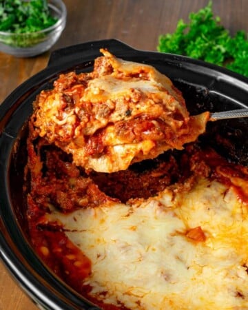 Slow Cooker Lasagna in a black crock with serving on a spatula.