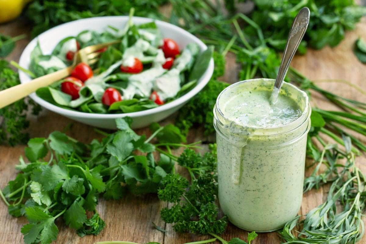 salad dressing in a jar with spoon, bowl of salad behind it.