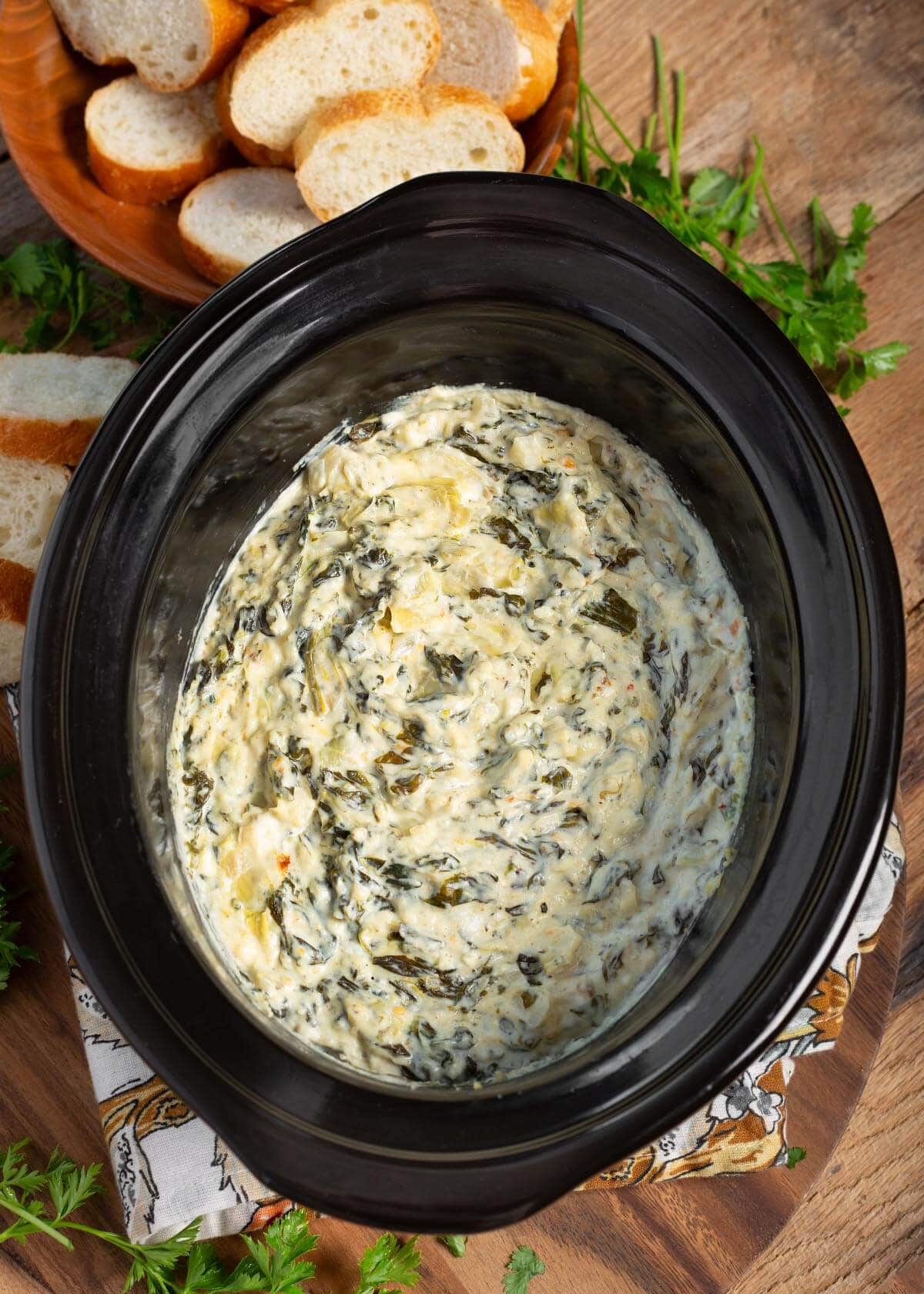 Slow Cooker Spinach Artichoke Dip in black crock with sliced baguette slices around it.