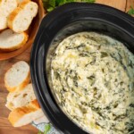 Slow Cooker Spinach Artichoke Dip in black crock with sliced baguette slices around it.